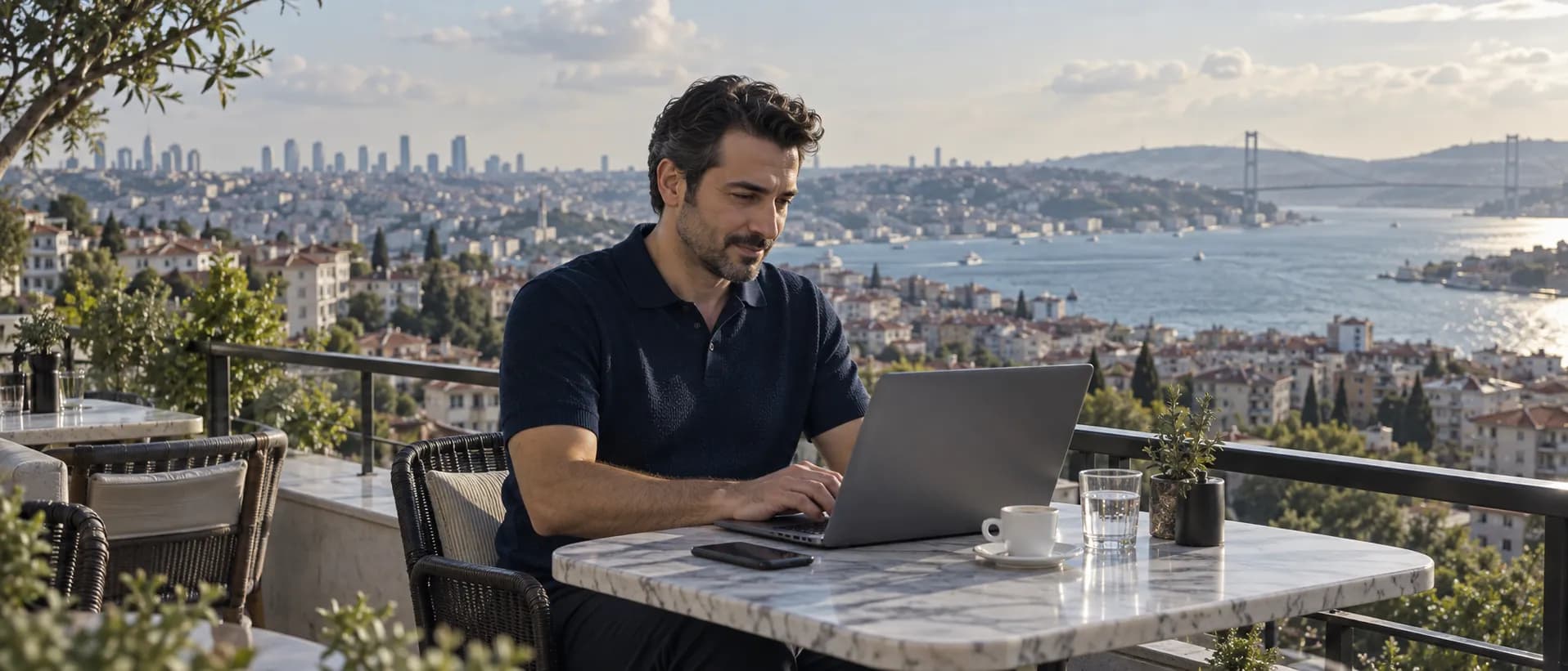 Terasta laptopla çalışan bir gayrimenkul danışmanı, arkasında İstanbul’un konutları ve Boğaz.